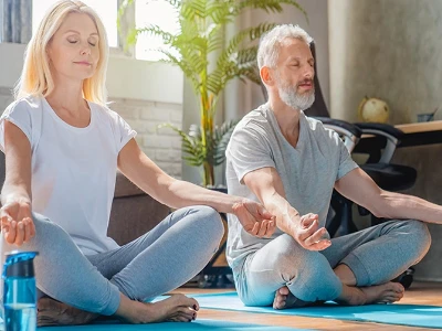 Couple mature assis en tailleur dans un salon, pratiquant la méditation ou la relaxation, les yeux fermés et les mains posées sur les genoux.