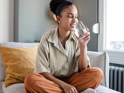 Femme assise sur un canapé, les yeux fermés, souriant en buvant un verre d’eau, dans un intérieur lumineux et apaisant.